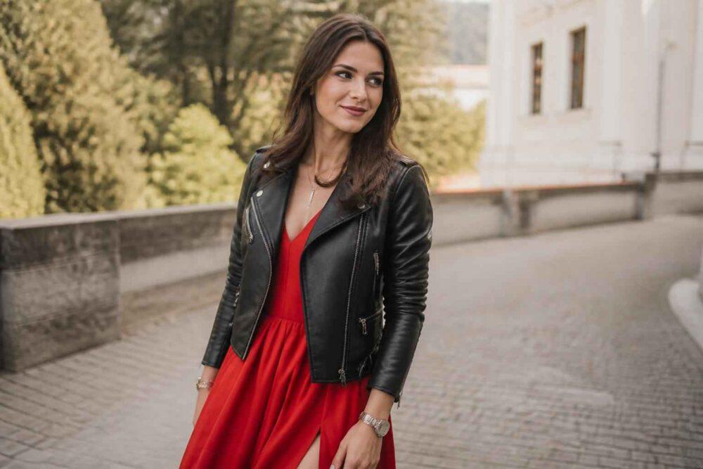 Kobieta w czerwonej sukience i czarnej skórzanej kurtce spaceruje przy eleganckim budynku.